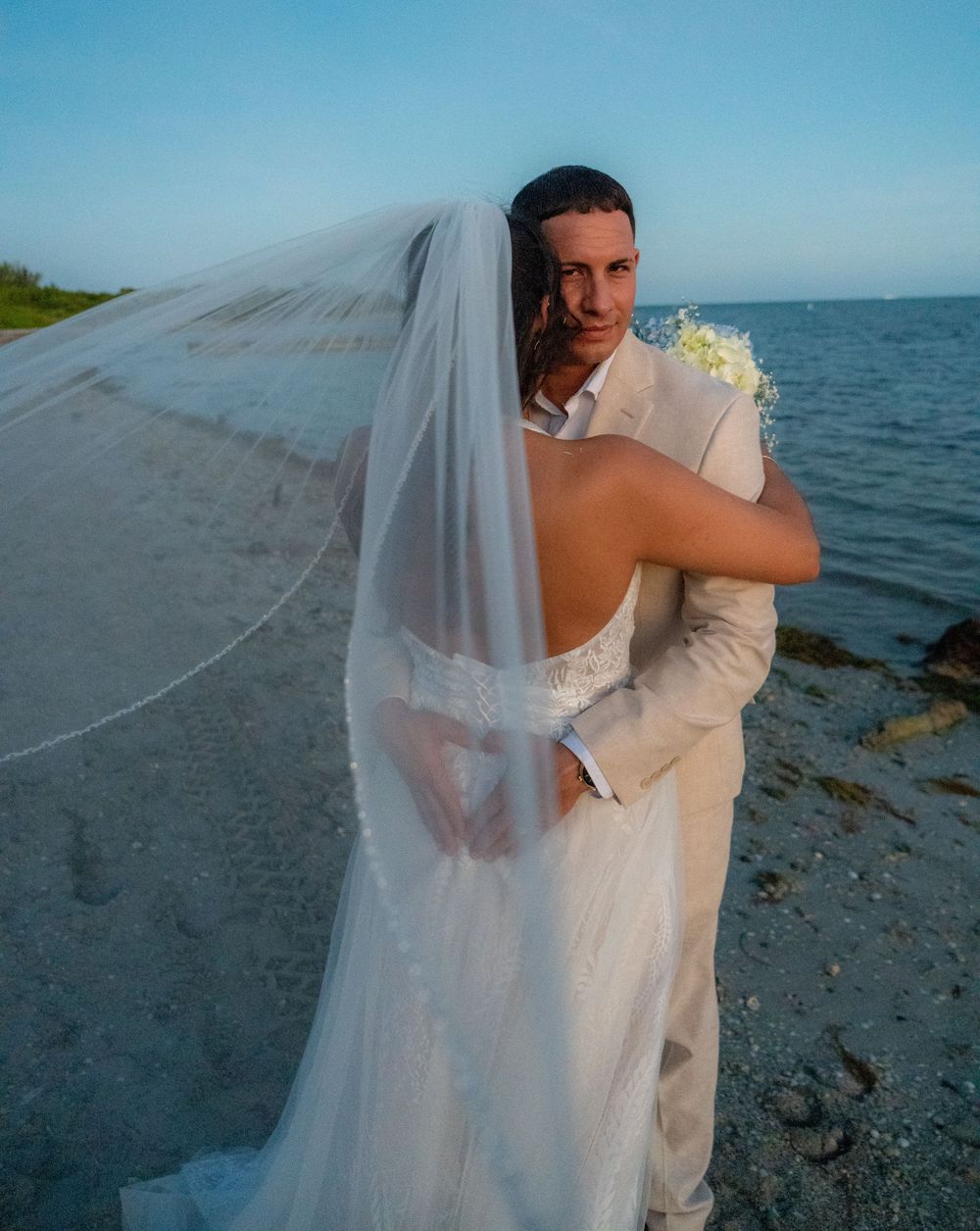 Groom embraces bride on beach at sunset as her long veil flows in the breeze.