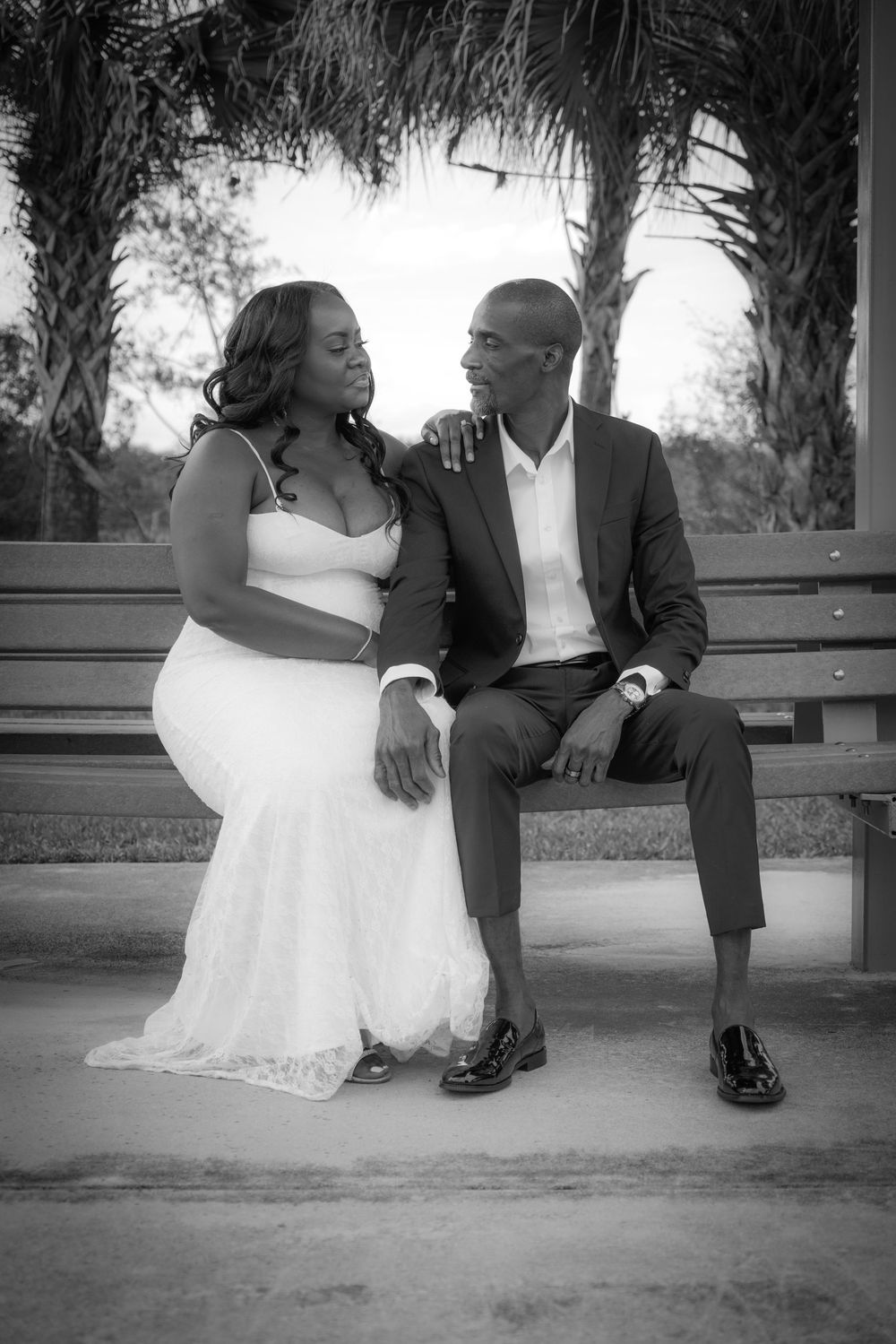 Black and white photo of bride and groom sitting on a bench under palm trees sharing a loving glance.
