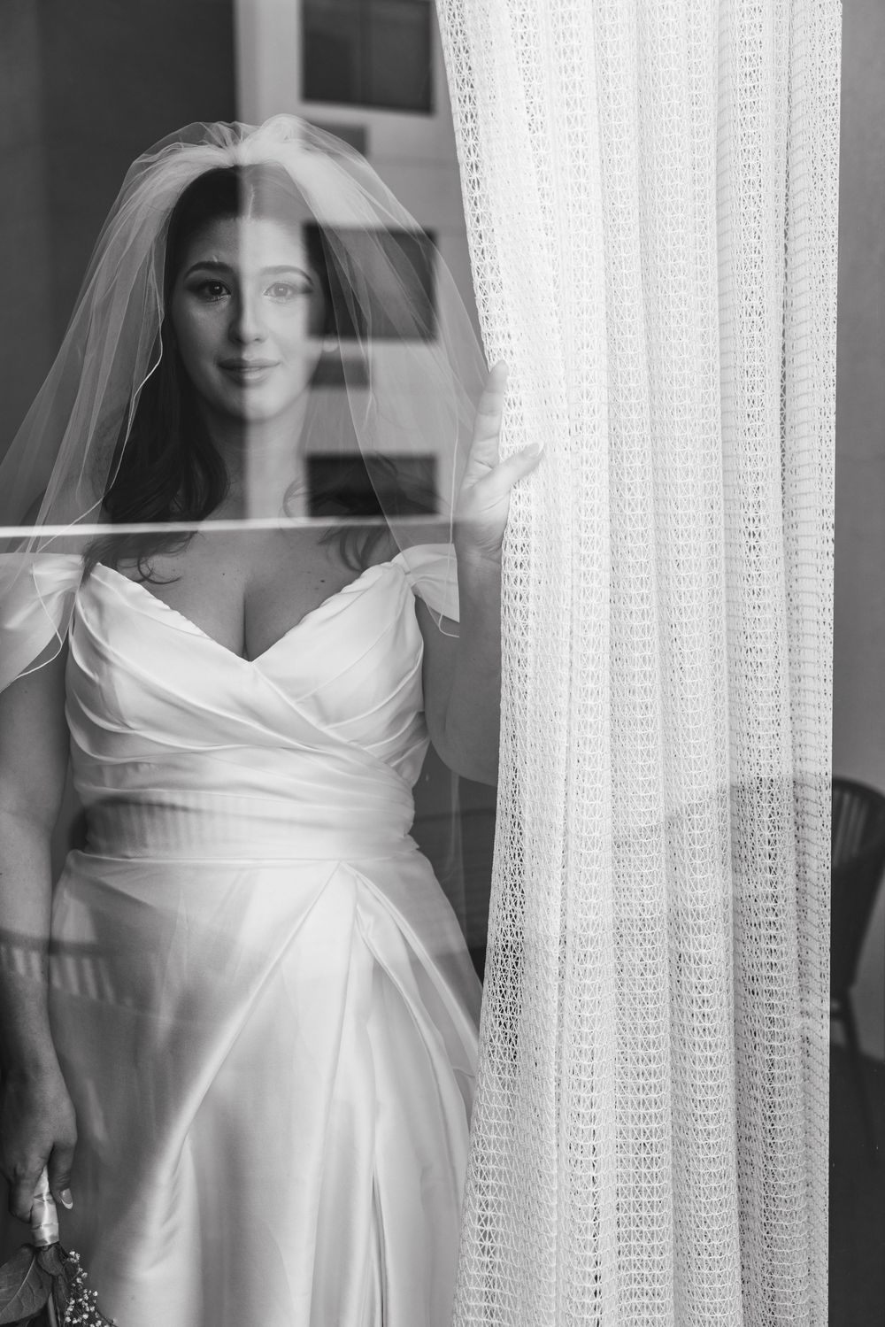 Elegant bride in satin off-shoulder gown and veil posing by window in black and white wedding portrait.