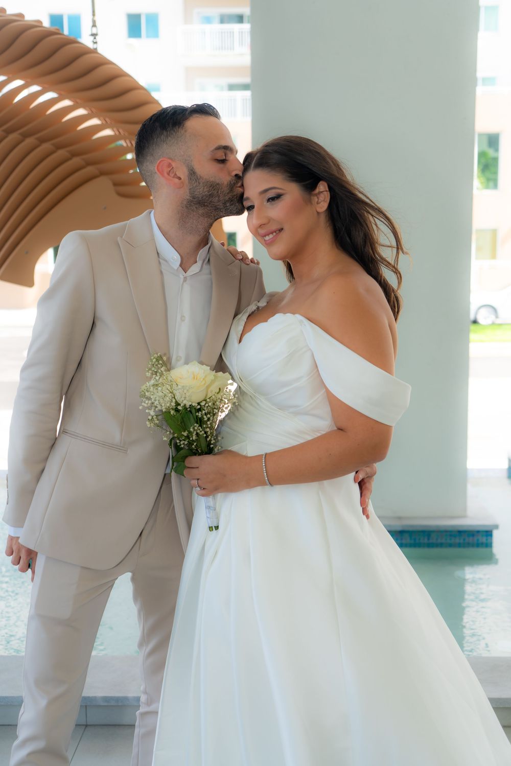 Groom in beige suit kissing bride in off-shoulder white gown holding white bouquet at outdoor wedding venue.