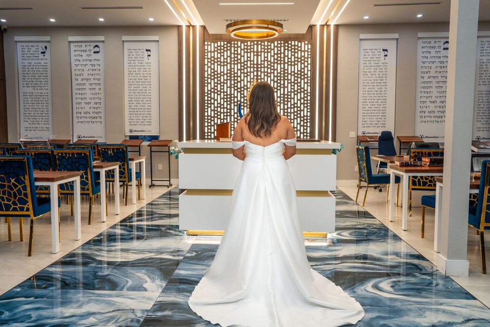 Bride in elegant white gown stands alone in beautifully decorated wedding venue interior.