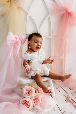 Happy baby girl in white lace dress sitting on white chair surrounded by pink tulle and floral decorations.