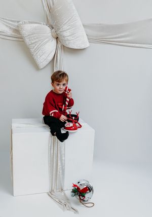 Young boy in red sweater sits on white box with candy cane, surrounded by giant Christmas bow decor.