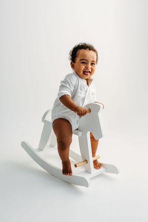 Smiling toddler in white outfit sitting on a white wooden rocking horse against a white background.