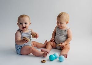 Two happy twin babies in matching gray rompers sitting together playing with wooden toys and blue balls on white background.
