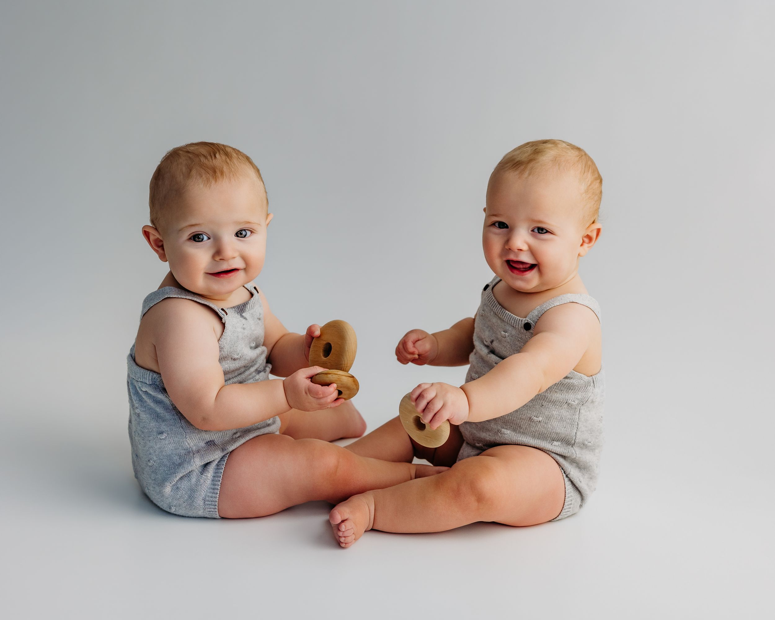 Two smiling baby twins in gray rompers sit together playing with wooden toys on a white background.