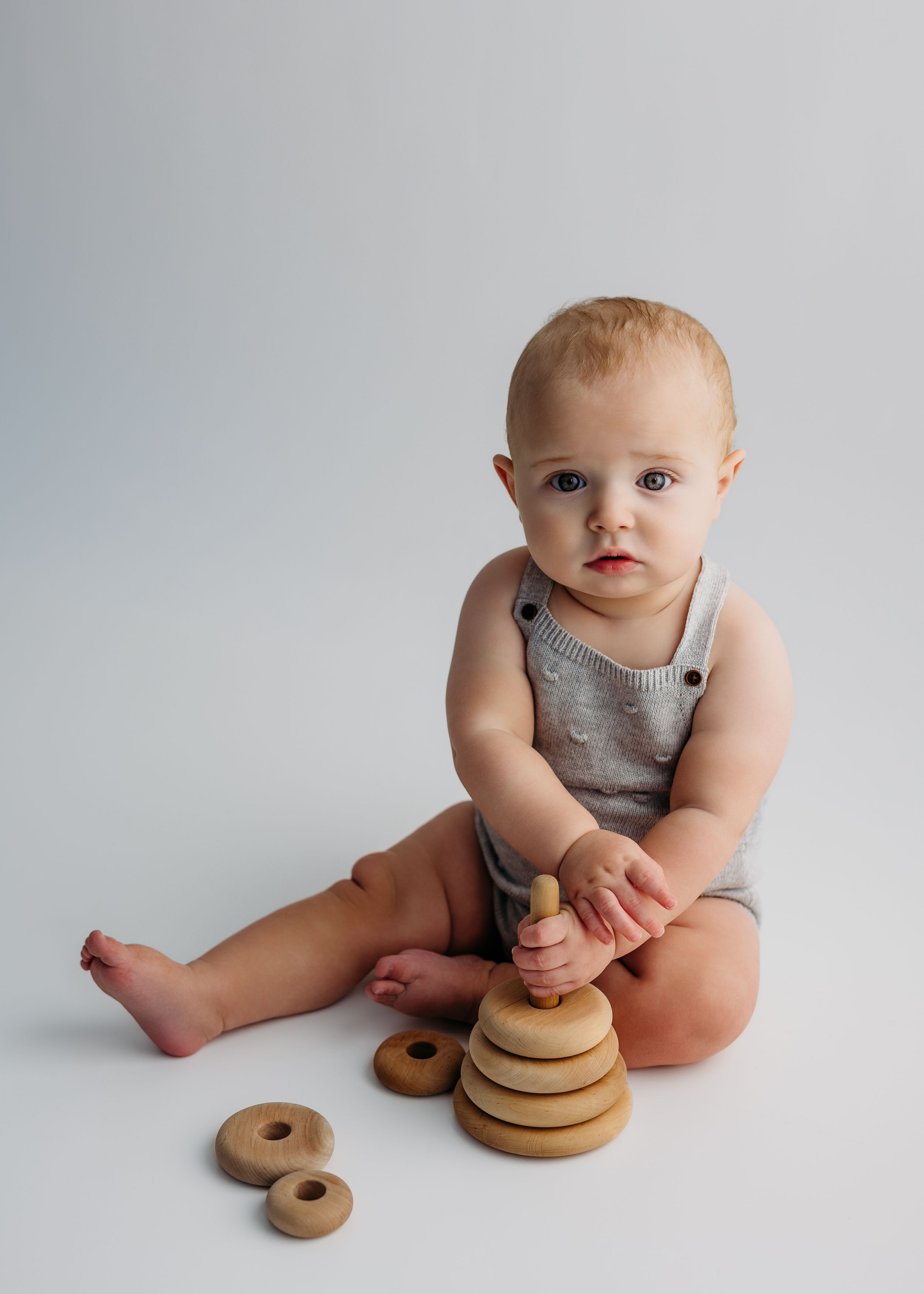 Baby in gray romper plays with wooden stacking ring toy while sitting on white background.