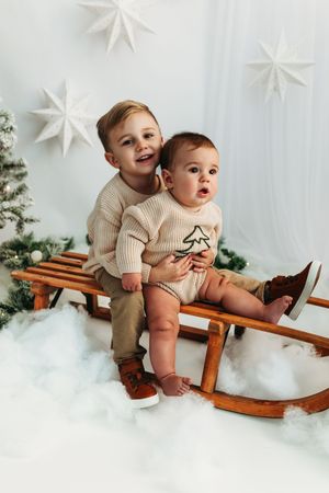 Two young boys in cream sweaters sit on a wooden sled with Christmas trees and star decorations.
