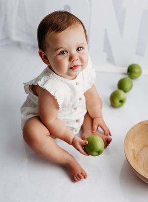 Adorable baby in white romper sitting on floor holding a green apple with more apples arranged nearby.