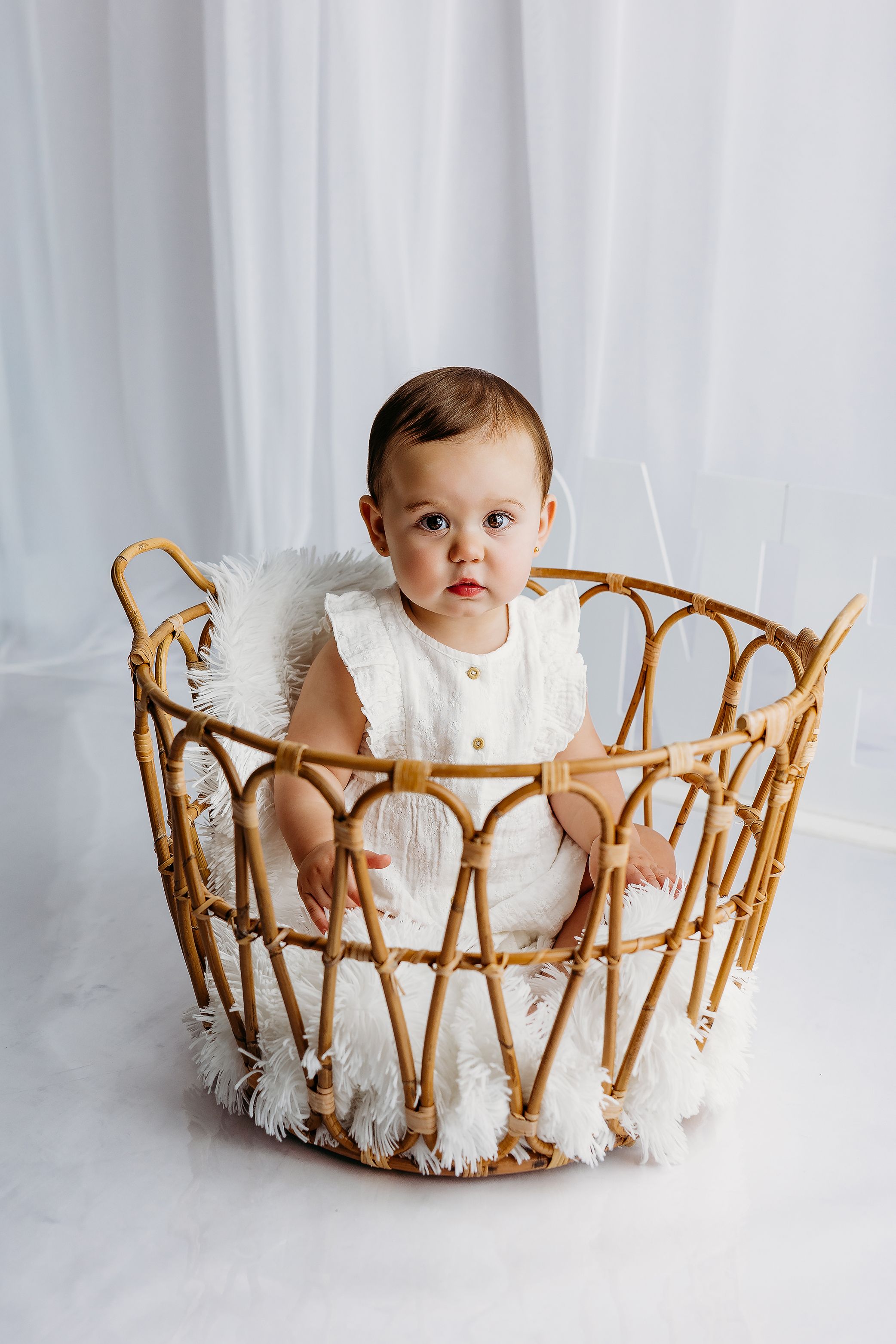 A cute baby in a white romper sits inside a decorative wicker basket with fluffy white lining.