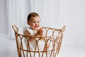A smiling baby in a white dress sits in a wicker basket, clapping hands against a soft white backdrop.
