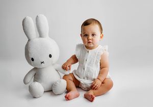 Baby in white romper sits beside a large Miffy bunny plush toy against a clean white background.