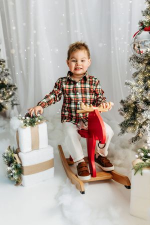 Young boy in plaid shirt rides red wooden rocking horse toy surrounded by Christmas trees and gifts.