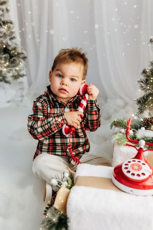 Toddler boy in plaid shirt holding candy cane phone near Christmas tree with vintage red rotary phone.