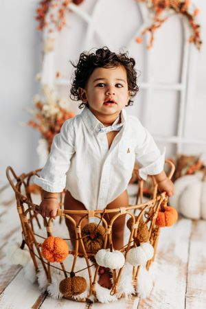 Toddler in white shirt standing in a wicker basket with pom-poms, fall decor background.