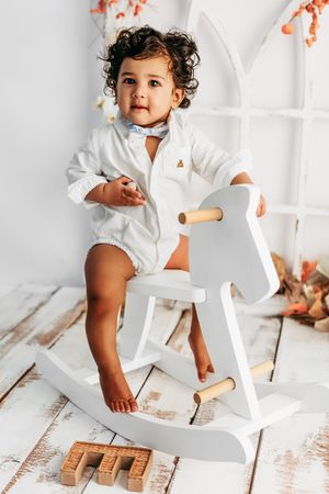 Curly-haired toddler in white shirt sitting on a white wooden rocking horse toy on rustic white floor.