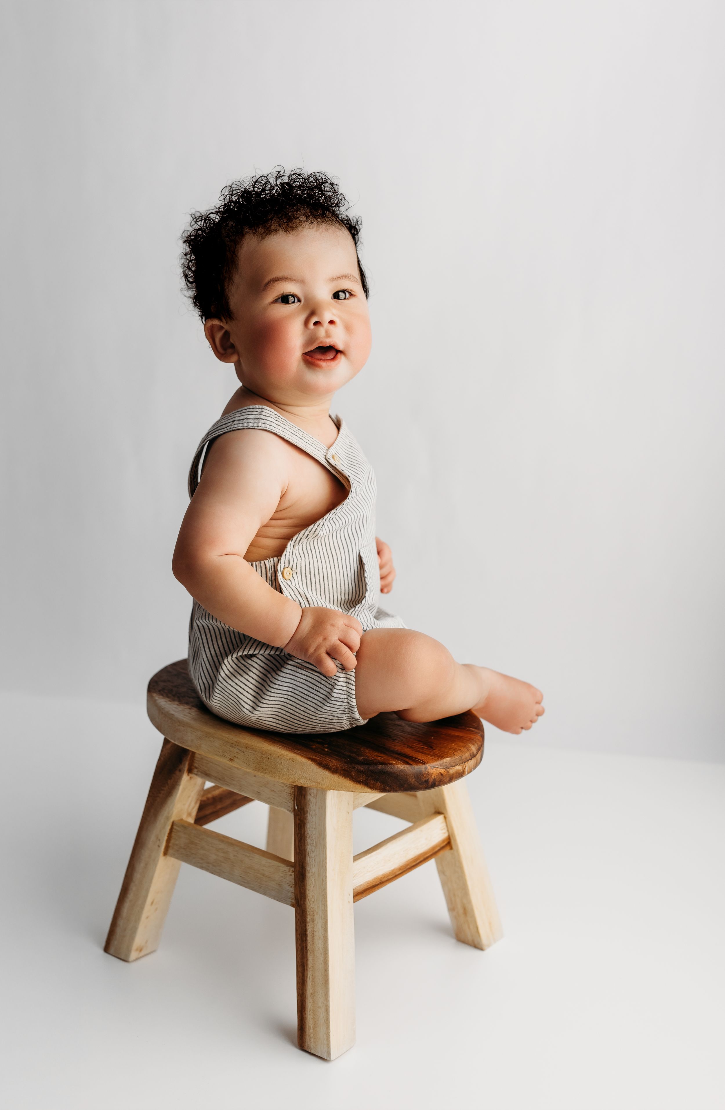 Smiling baby in striped romper sitting on a small wooden stool against a white background.
