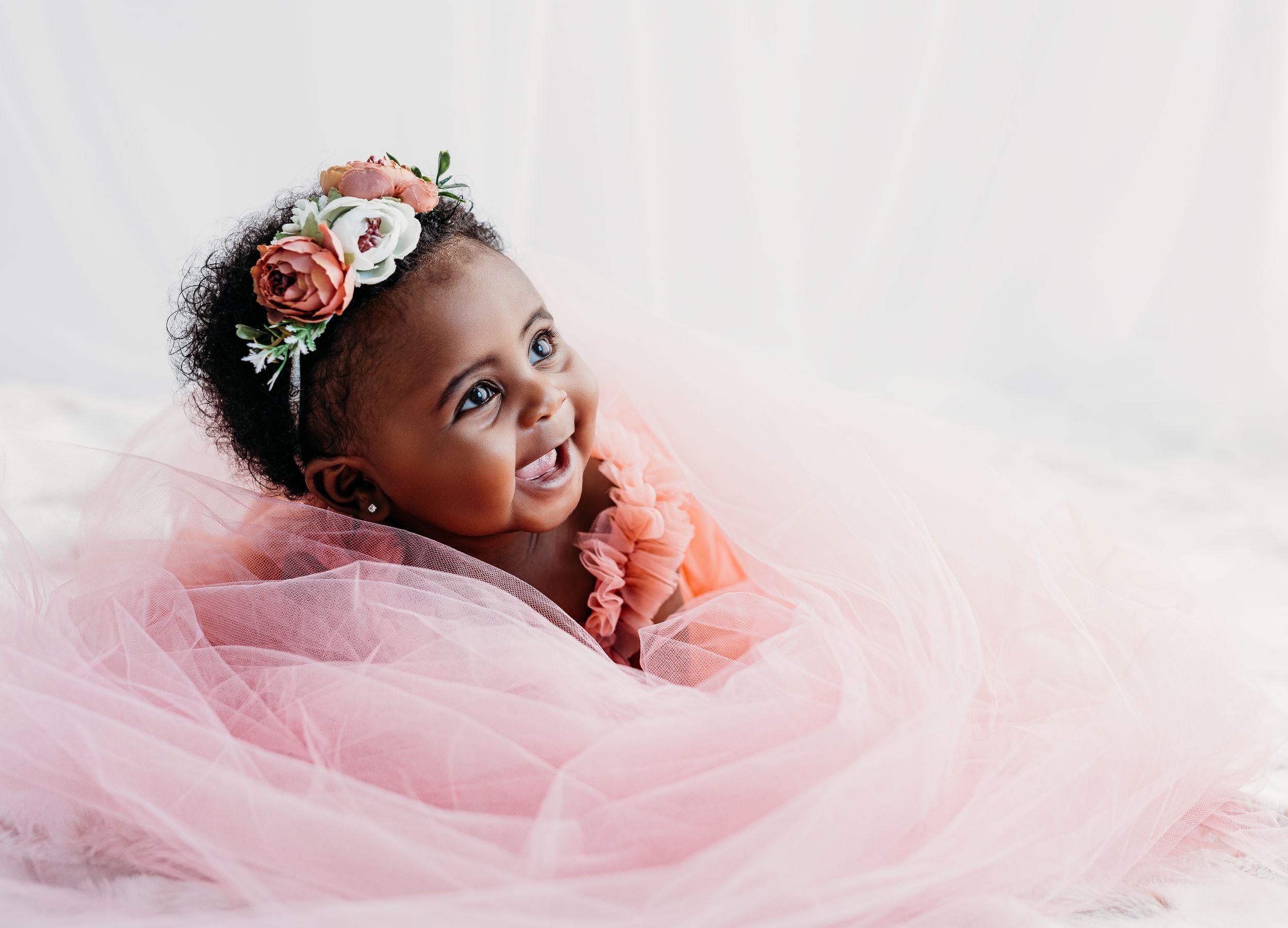Adorable baby girl in pink tulle dress and floral headband smiling during professional photo shoot.