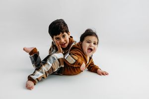 Two young children in brown and plaid outfits laugh and pose together against a white background.