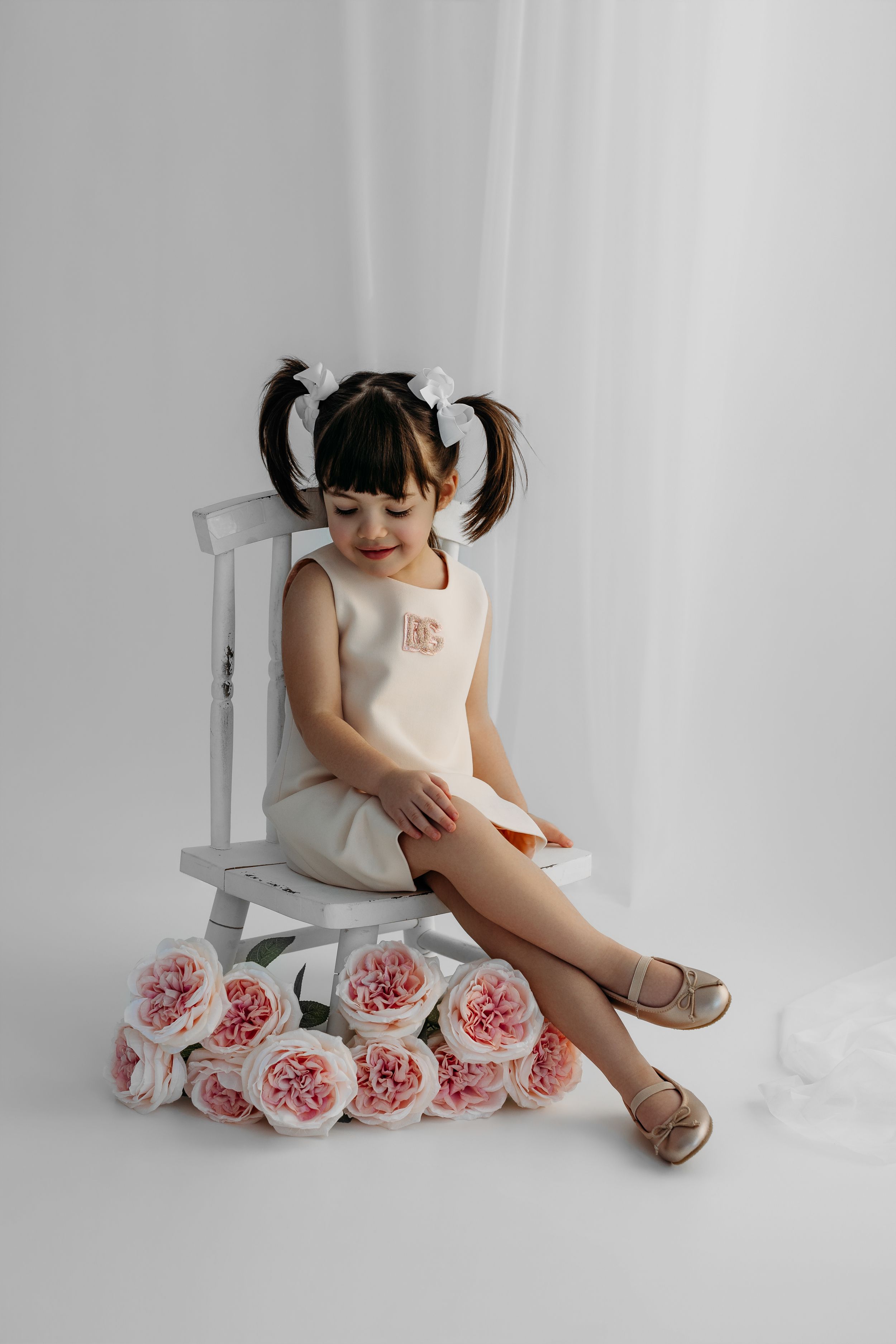 Young girl in cream dress with pigtails sits on white chair surrounded by pink roses in a bright studio setting.
