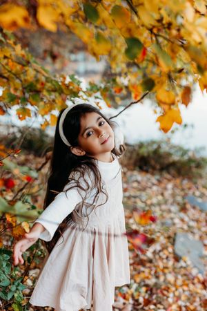 Young girl in white dress playing under colorful autumn leaves by a lake, wearing ear muffs.