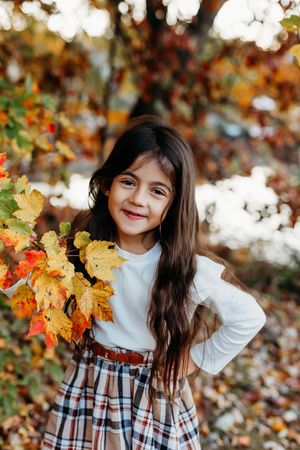 A smiling young girl with long dark hair holds colorful autumn leaves, wearing a white top and plaid skirt outdoors.