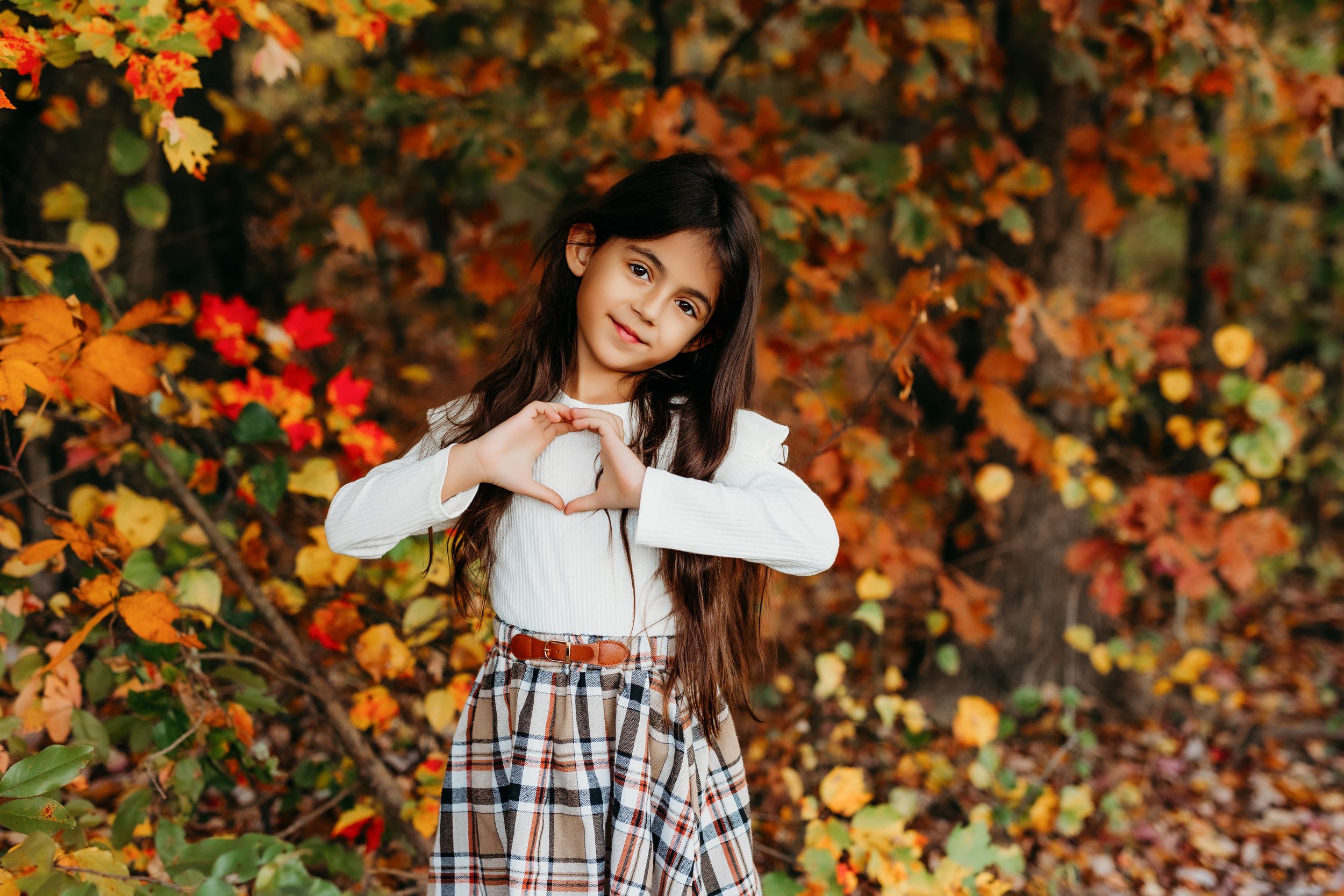 A young girl in a plaid skirt makes a heart shape with her hands outdoors among vibrant autumn foliage.