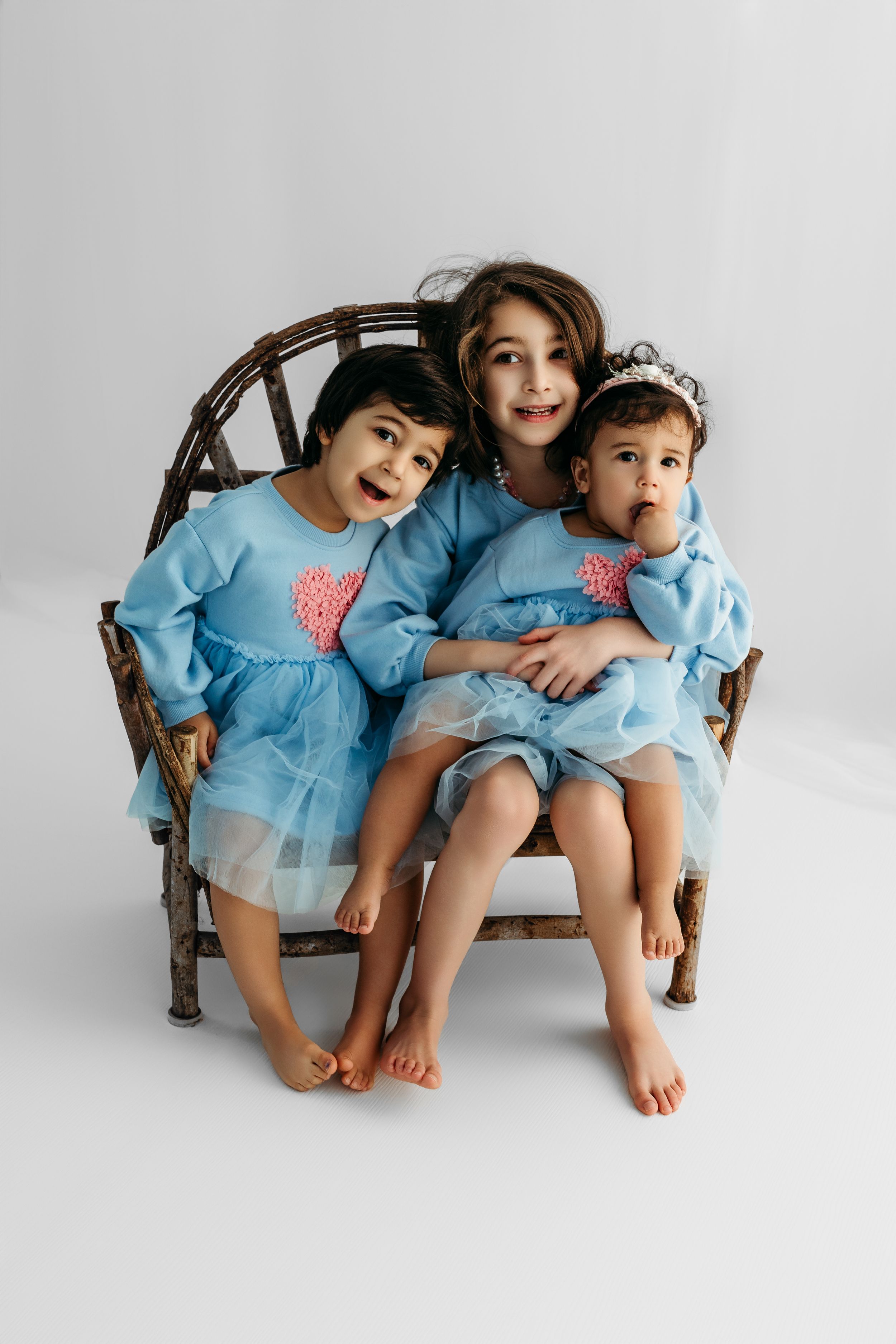 Three young sisters in matching blue dresses with pink hearts sit together on a rustic twig bench for a studio photo.