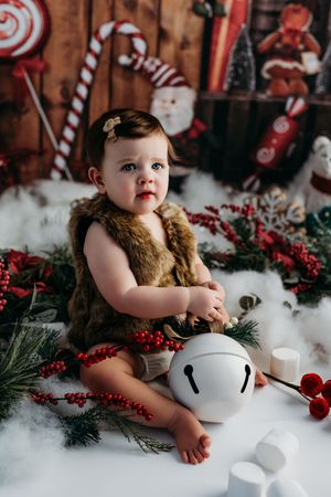 Adorable baby girl in fur vest sits among Christmas decorations, bells, and red berries in a festive holiday photo session.