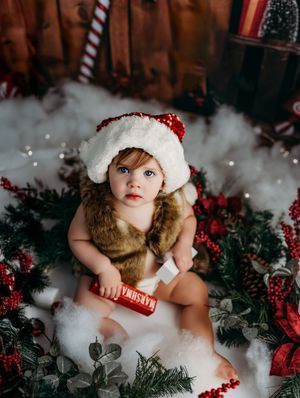 Adorable baby in Santa hat and fur vest sits among Christmas greenery and red berries holding a candy bar.