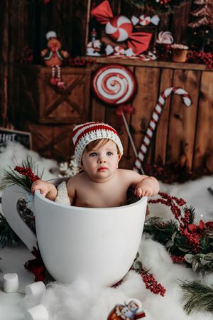Baby in striped knit hat sits in giant white mug amid Christmas candy cane decorations and festive holiday backdrop.