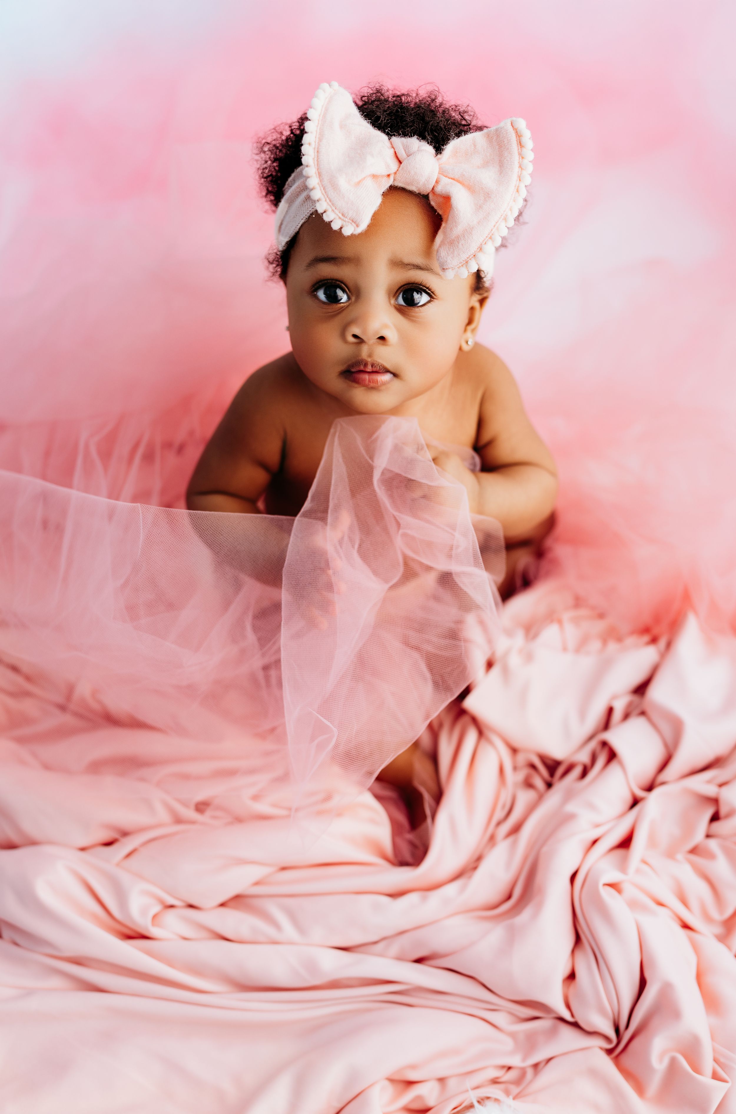Adorable baby girl in pink tulle skirt and bow headband sitting on soft pink fabric in a dreamy photoshoot.
