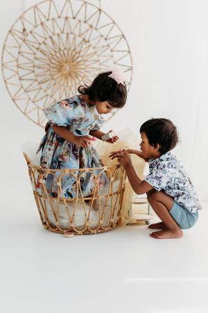 Two young children in floral outfits playing with a wicker basket in a bright white studio setting.