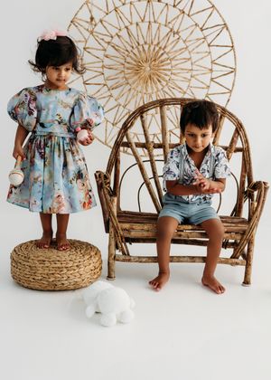 Two toddlers in floral outfits pose with a rustic wooden chair and wicker stool against a white background.