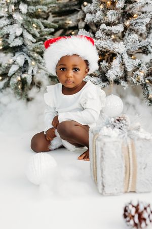 Adorable Black baby in Santa hat and white outfit sits by snow-flocked Christmas tree with silver gifts.