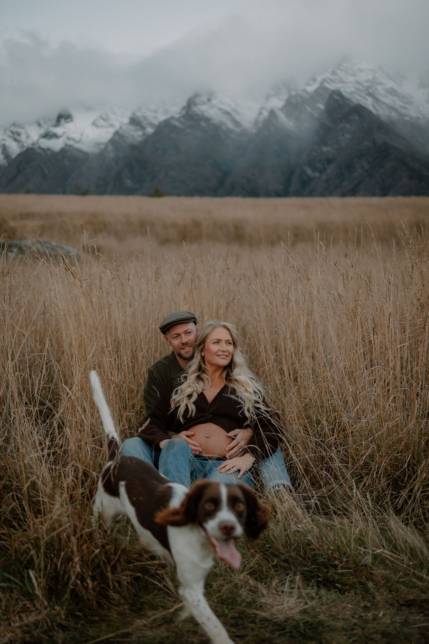 Pregnant couple sits in tall grass field with snow-capped mountains backdrop and brown and white dog in foreground.