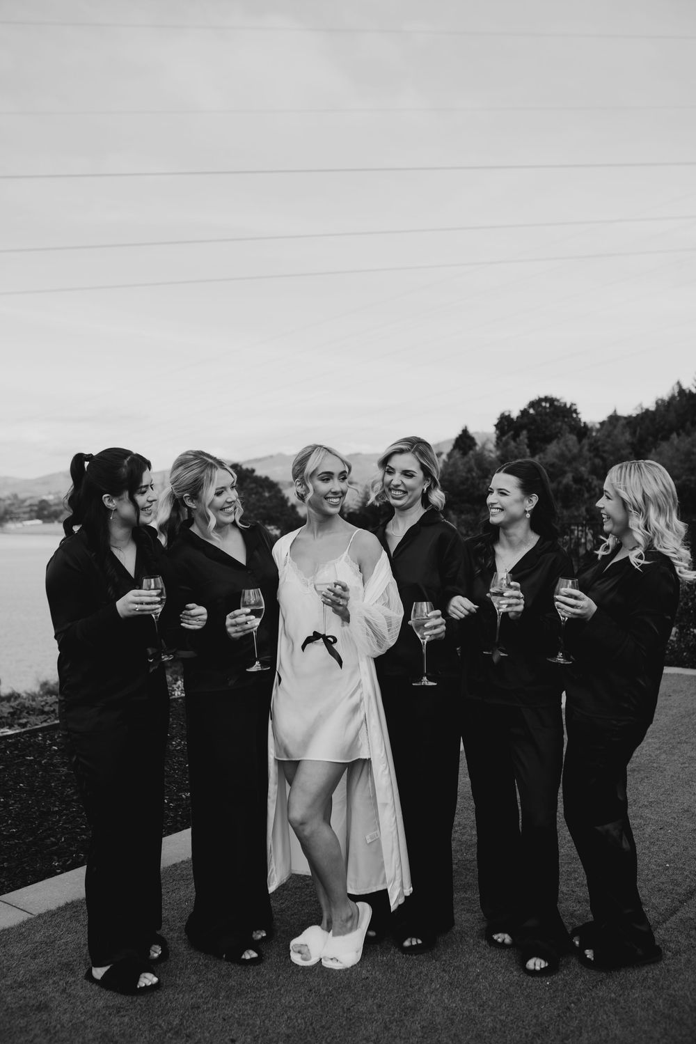 Bride in a white robe celebrates with bridesmaids in black pajamas holding champagne glasses outdoors.