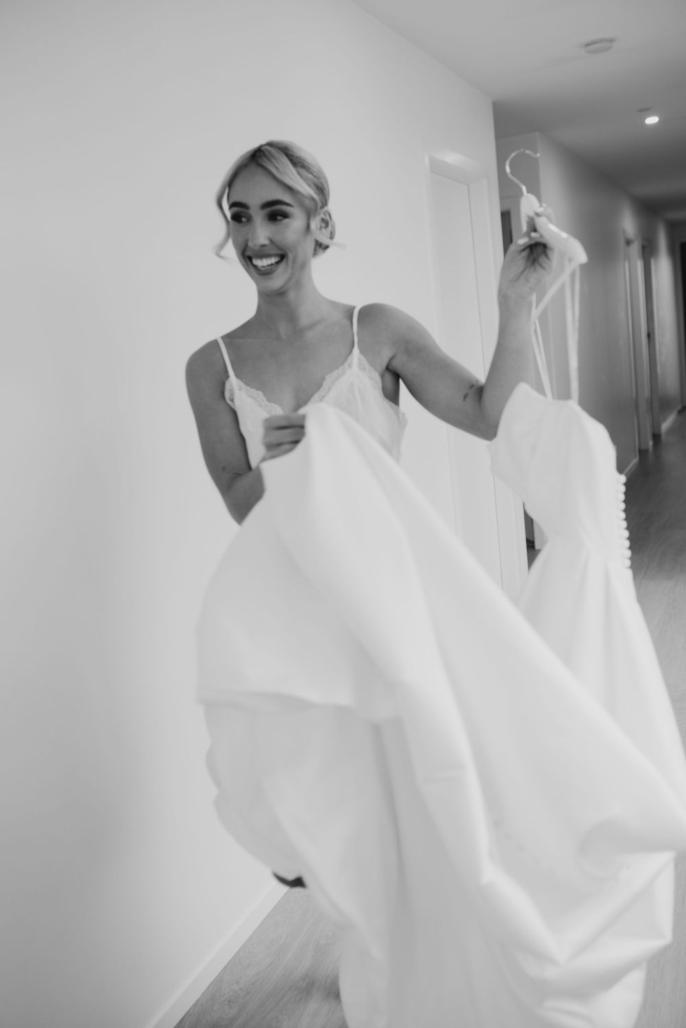 Smiling bride in a slip dress holds her wedding gown on a hanger in a bright hallway before getting dressed.