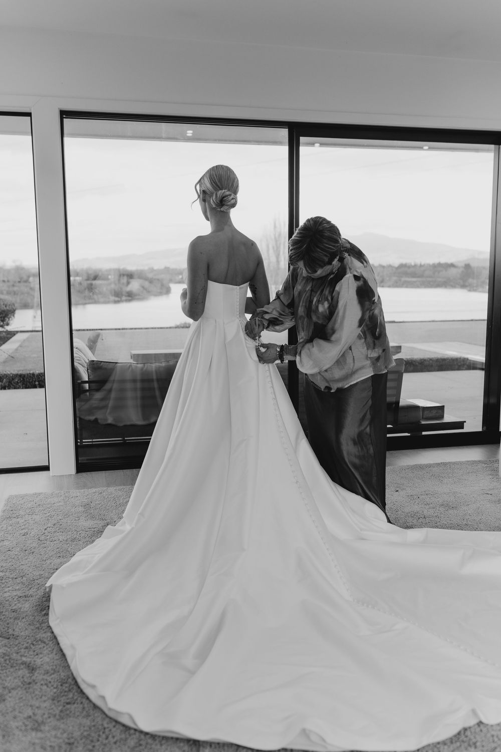 Black and white photo of a bride in a strapless gown being buttoned up by her bridesmaid near a window.