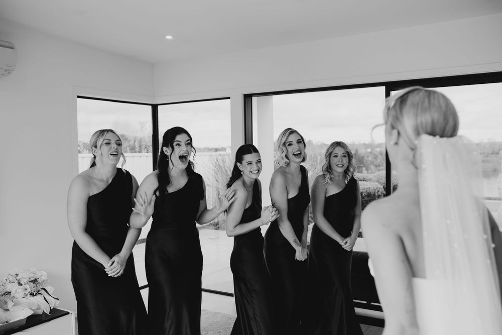 Bridesmaids in black dresses react with joy seeing the bride in her wedding gown for the first time.