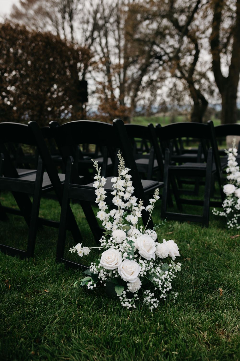 White roses and baby's breath floral arrangement beside black folding chairs at an outdoor wedding ceremony.