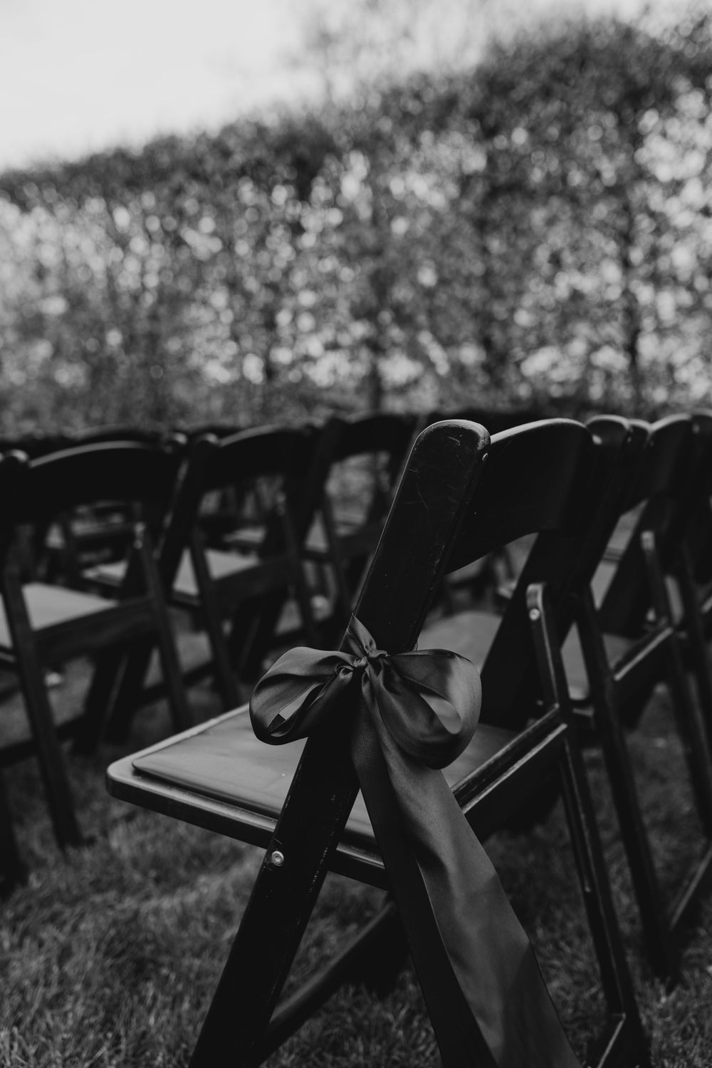 Black and white photo of wooden folding chairs with satin ribbon bows at an outdoor wedding ceremony.
