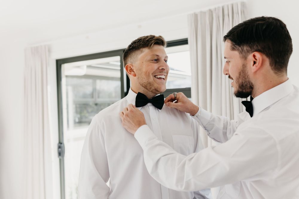 Smiling groom has his black bow tie adjusted by his best man while getting ready for the wedding.