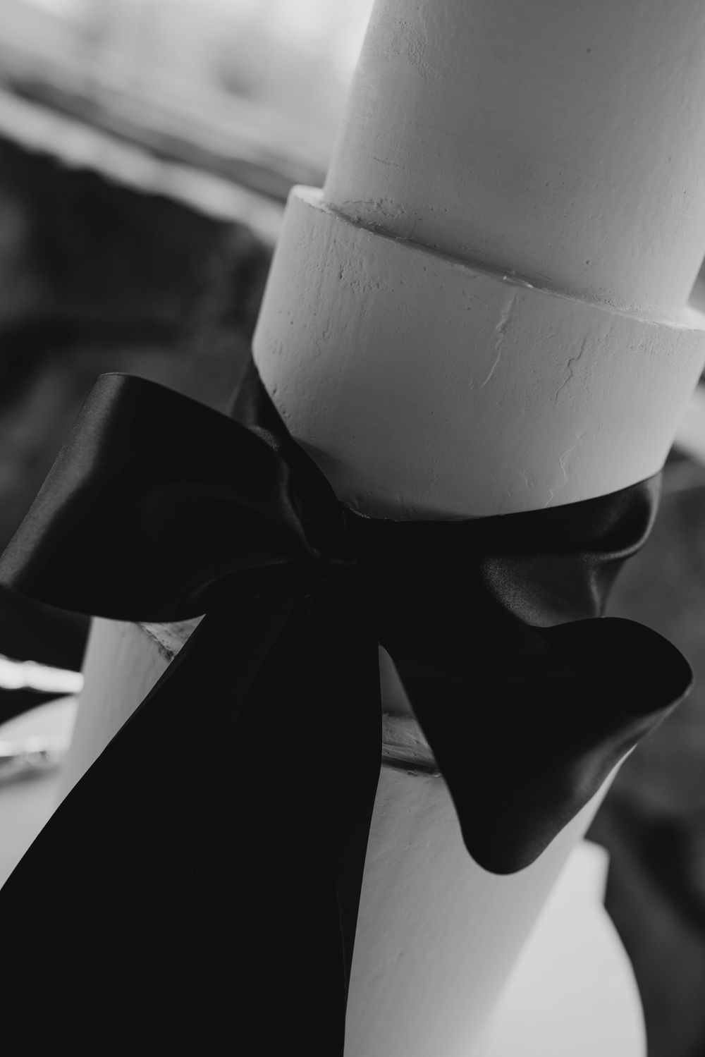 Close-up black and white photo of a white wedding cake tier adorned with a large black satin ribbon bow.