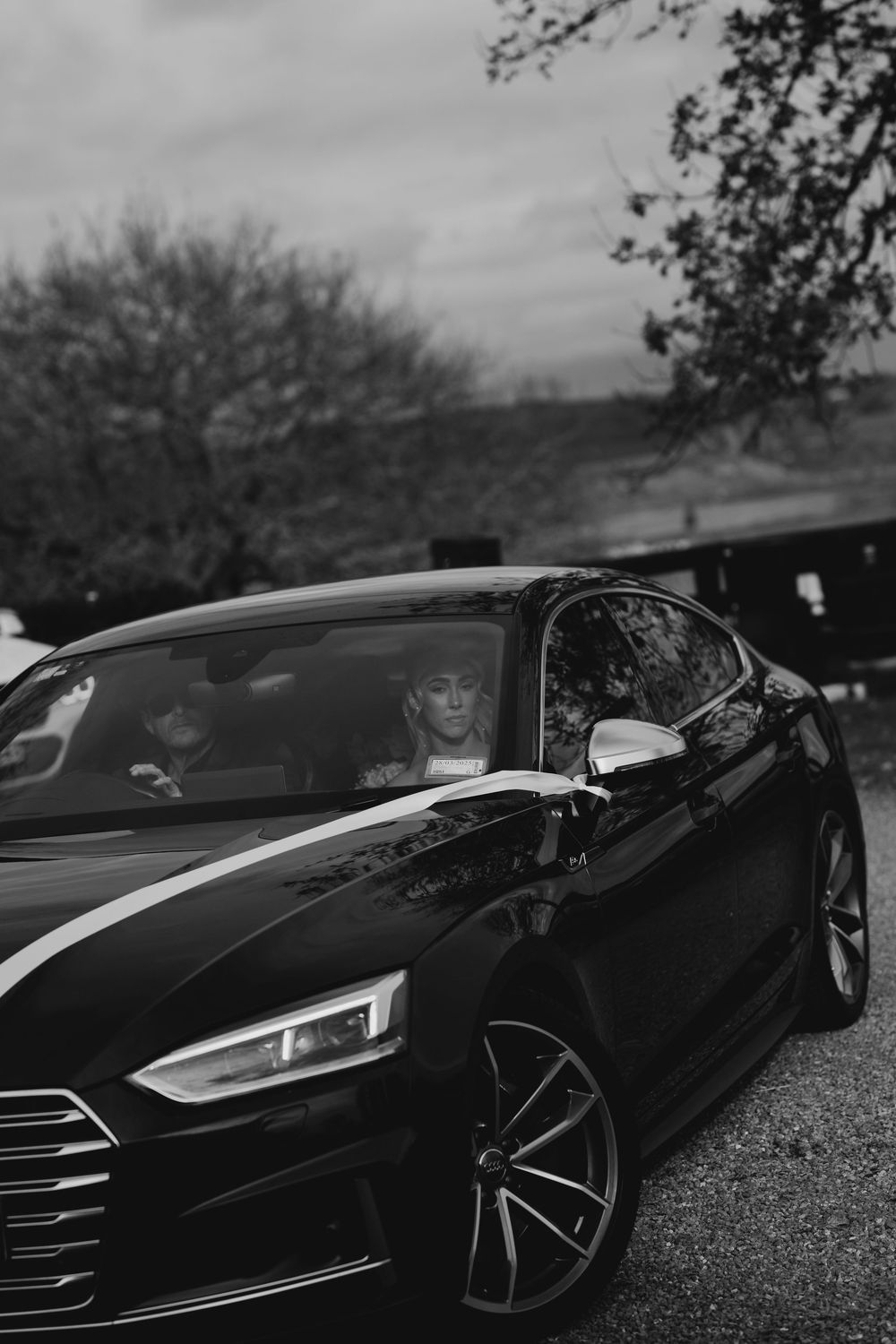 Black and white photo of bride seated inside a sleek black Audi luxury car on wedding day.