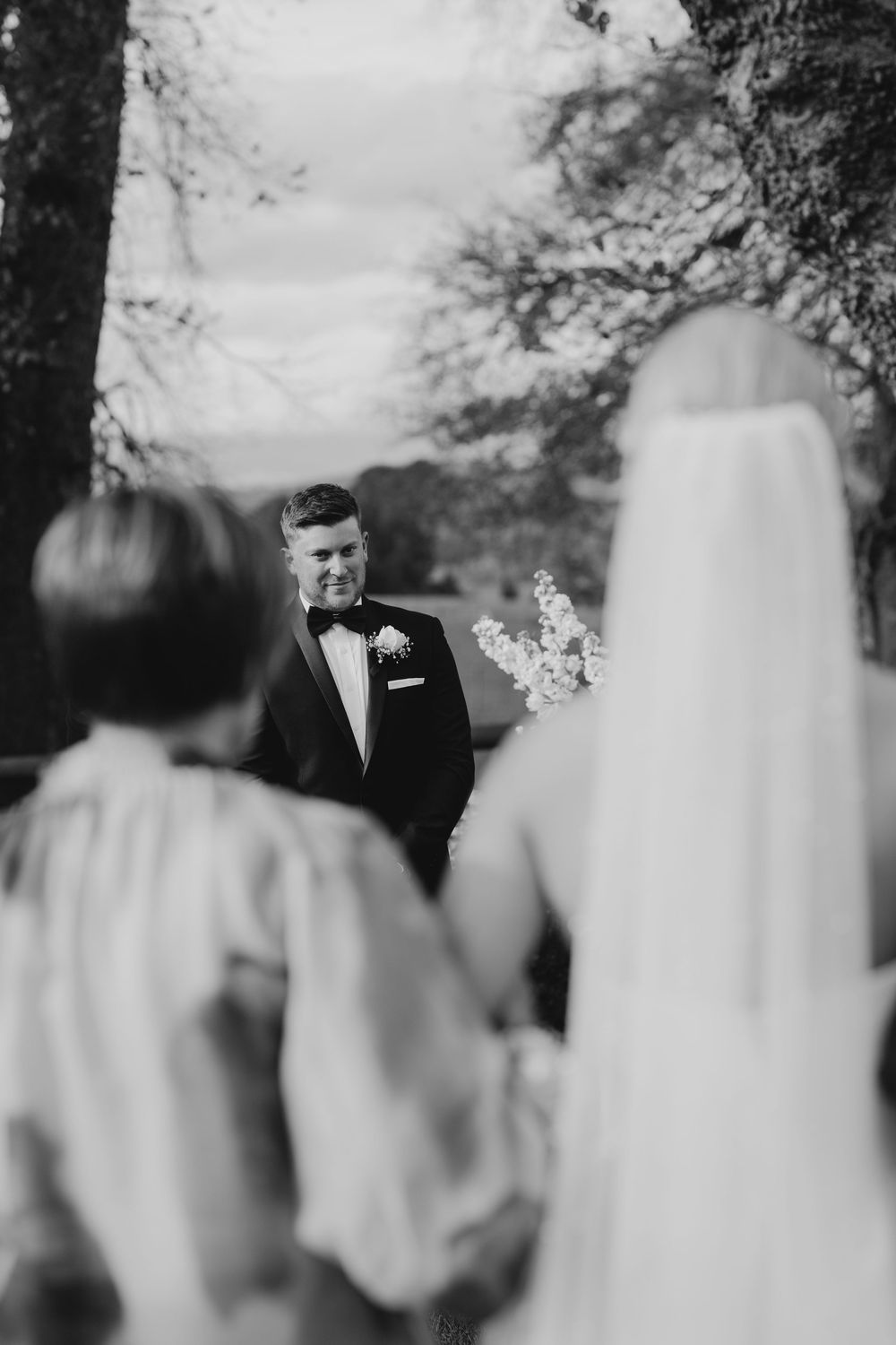 Black and white photo of emotional groom in tuxedo watching bride approach down the aisle at outdoor ceremony.