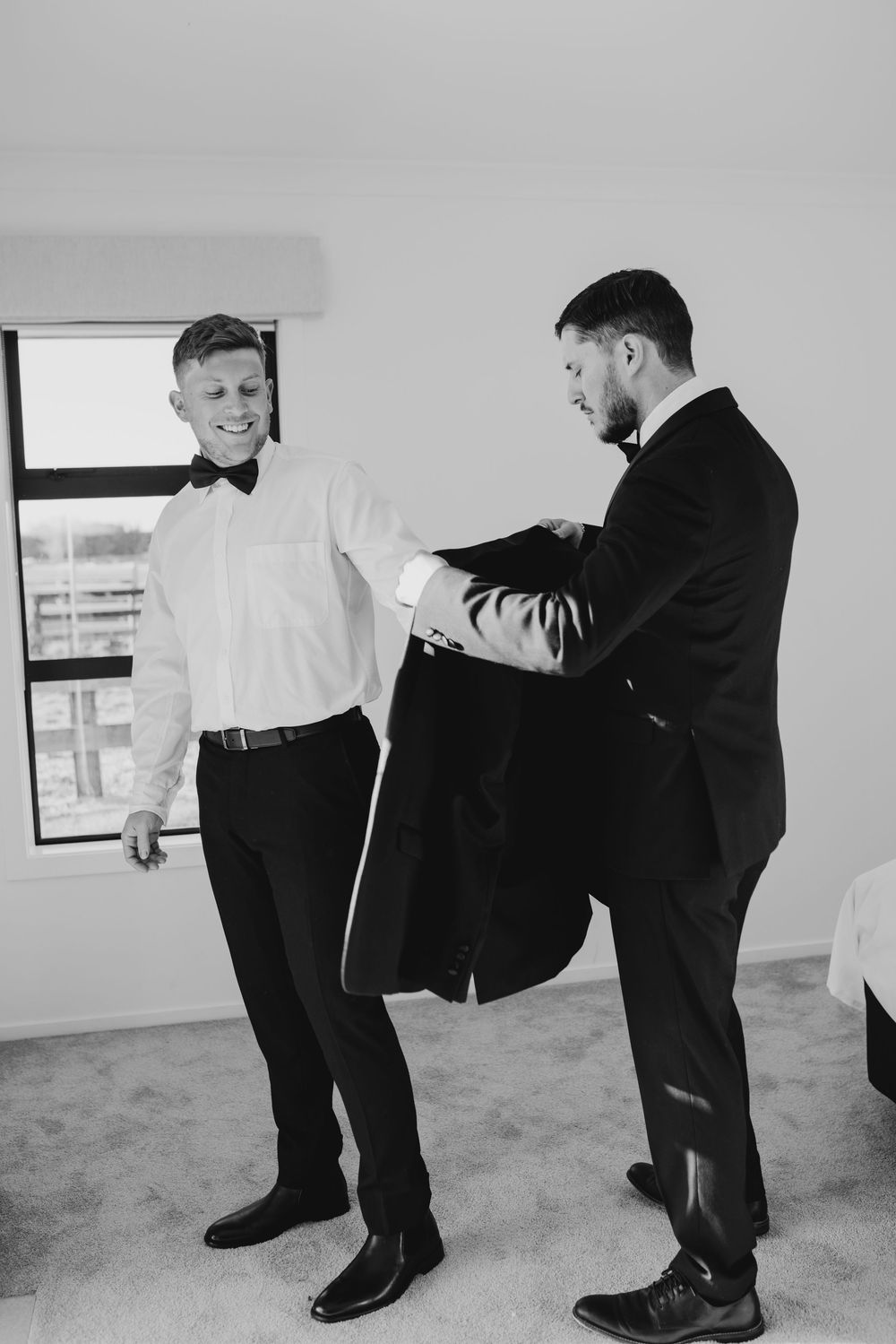 Black and white photo of a best man helping the groom into his suit jacket before the wedding ceremony.