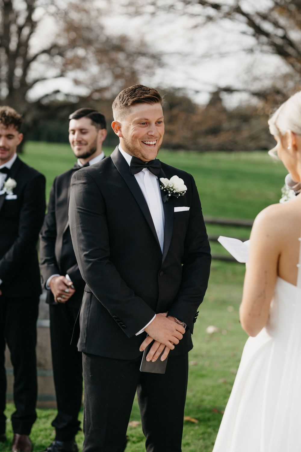 Smiling groom in black tuxedo with white boutonniere listens to bride read vows at outdoor wedding ceremony.