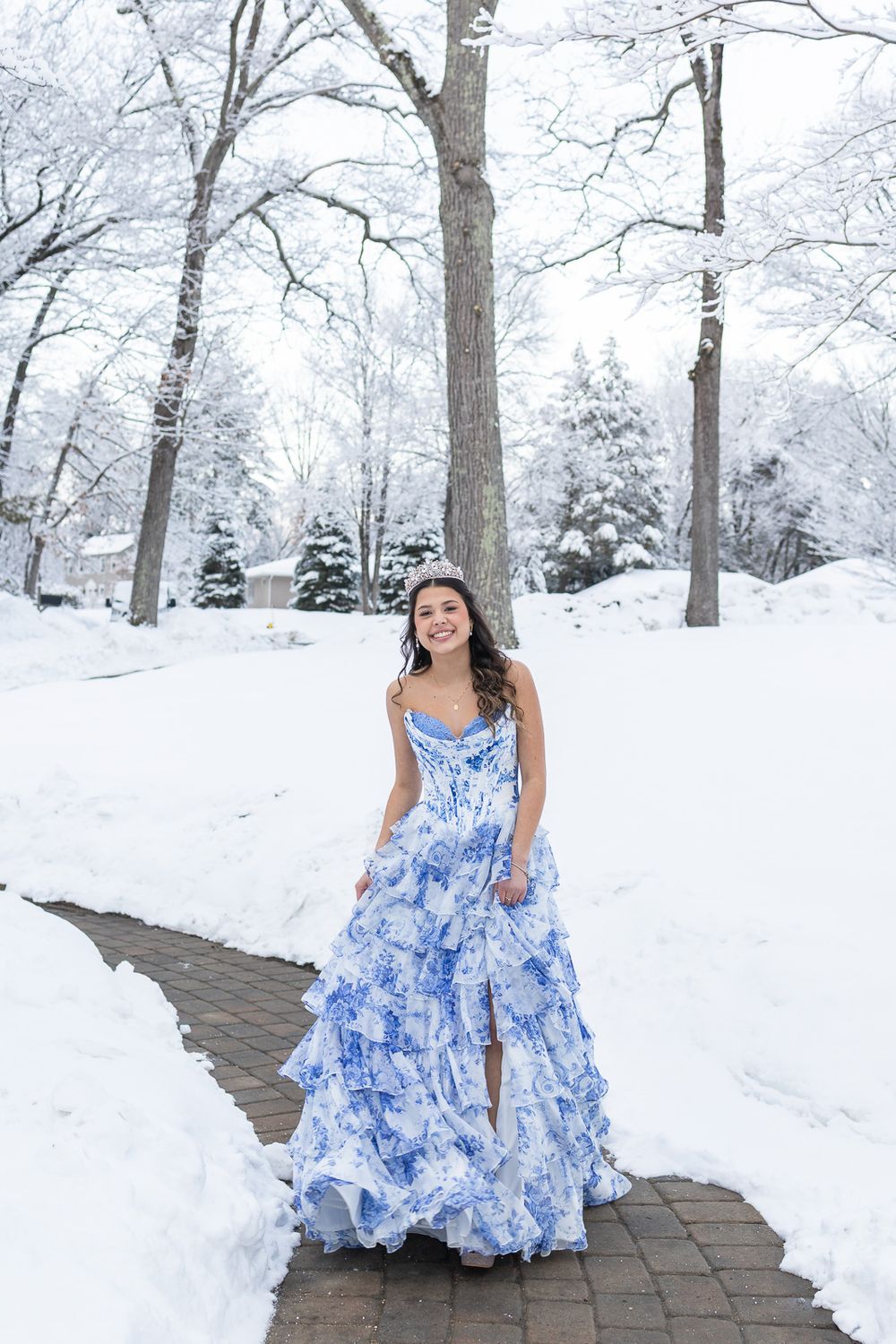 Young woman in blue floral ball gown wearing tiara poses on snowy brick path surrounded by snow-covered trees.