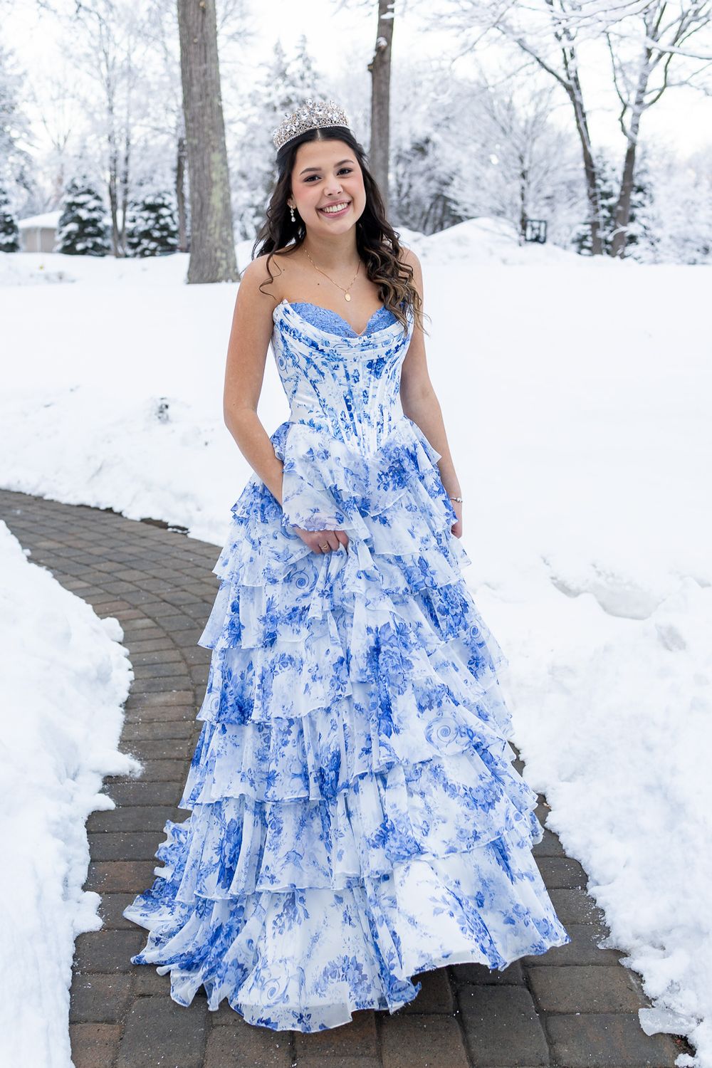 Young woman in blue floral tiered prom gown with tiara poses on snowy brick path outdoors.